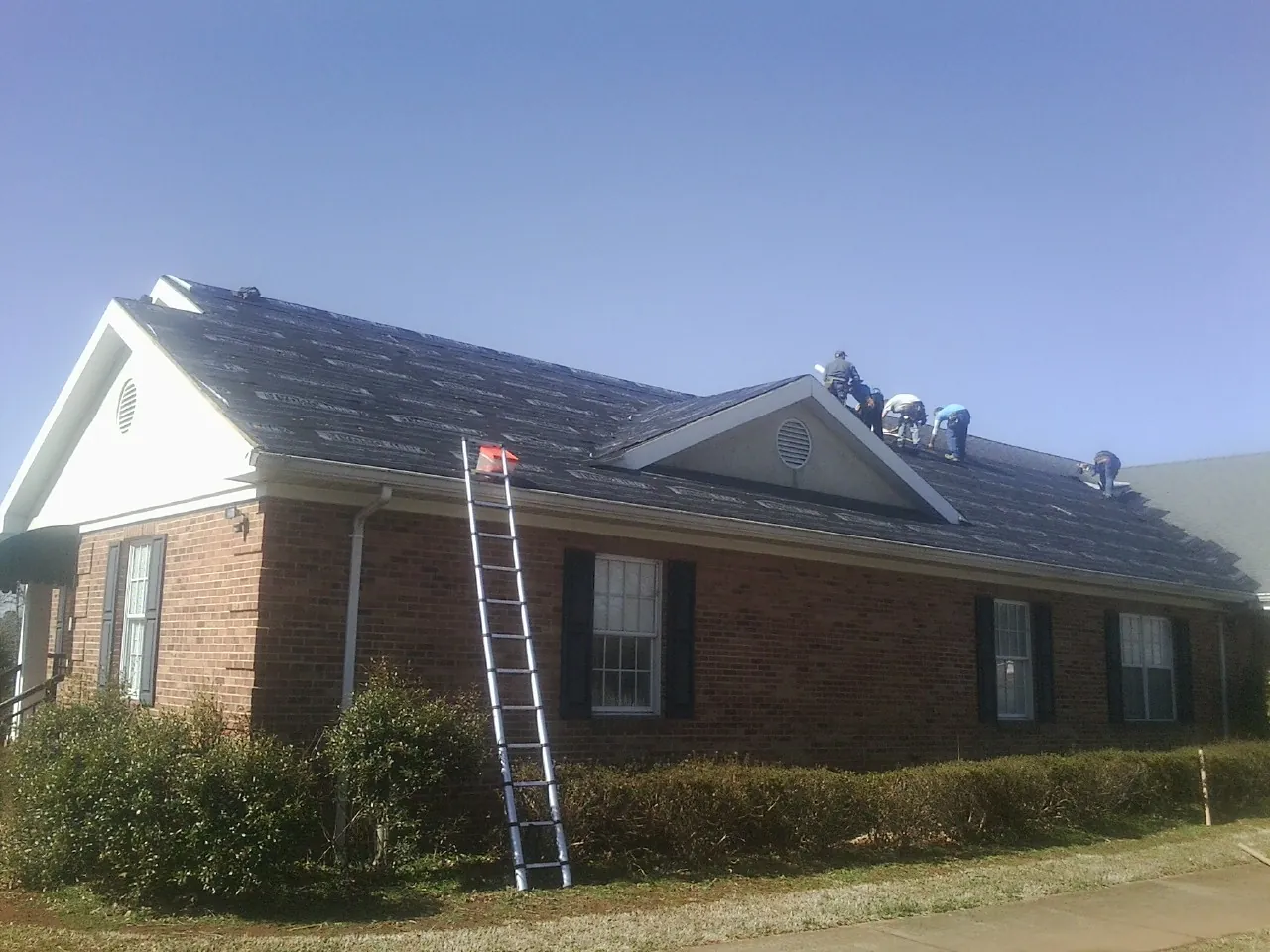 Crew tearing shingles off a brick ranch roof with a ladder set — Metal Roof Repair work by our crew in Nye