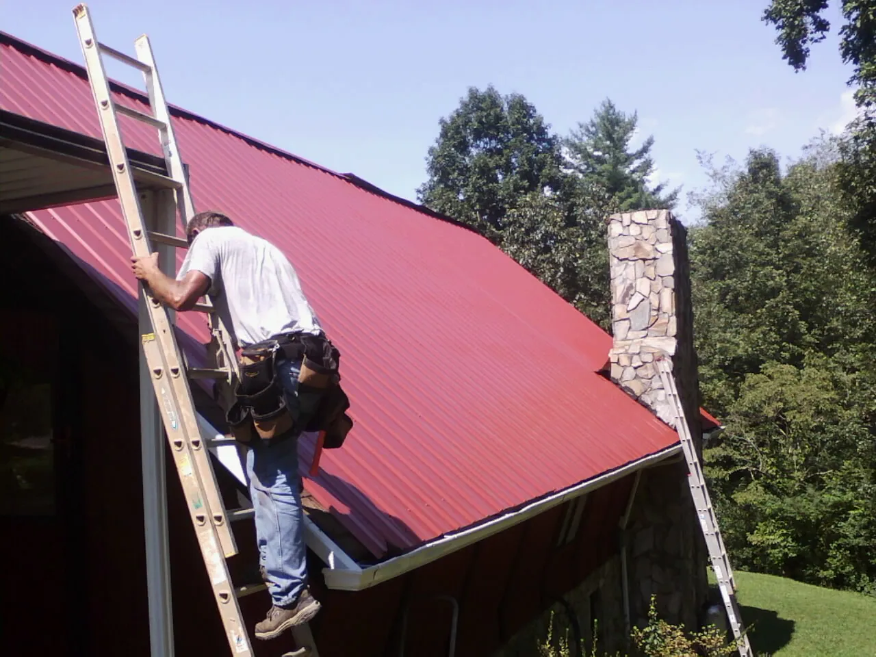 Roofer on a ladder beside a red metal roof and stone chimney — Soffit & Fascia Repair by Roofing Brothers in Nye