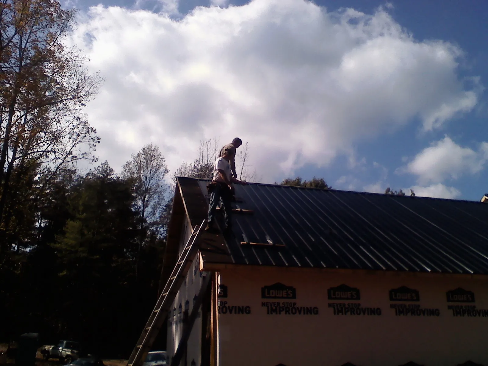 Roofer fastening dark metal panels high against a cloudy sky, from a Roof Inspection job in Nye